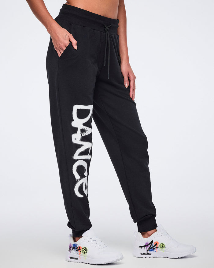 Female model wearing black joggers with bold white “DANCE” lettering down her right leg and white graffiti‑accented sneakers against a white background.