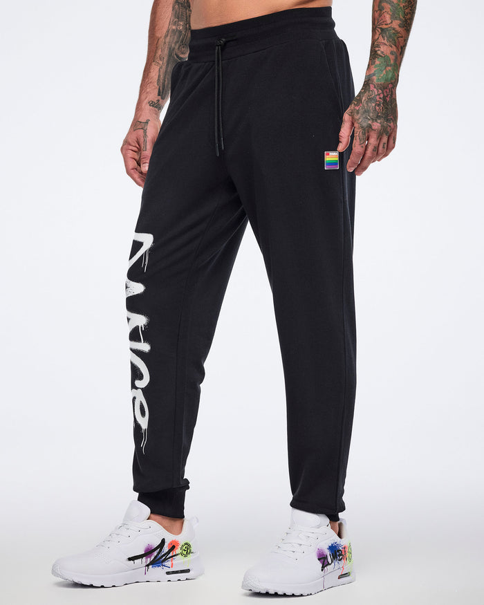 Male model wearing black jogger pants with showing a small rainbow long on the upper left leg and partially visible "DANCE" down the right one. The model is wearing white shoe with graffiti-style print all shown on a white background.