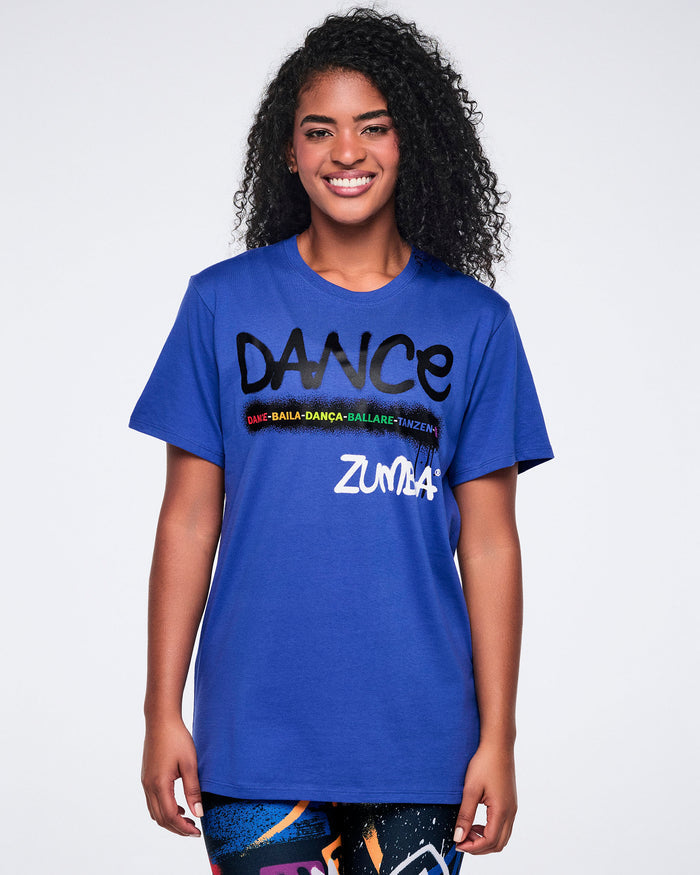 Female model wearing a blue Zumba t-shirt with 'DANCE' printed in black and 'Zumba' in white, paired with colorful leggings against a white background.