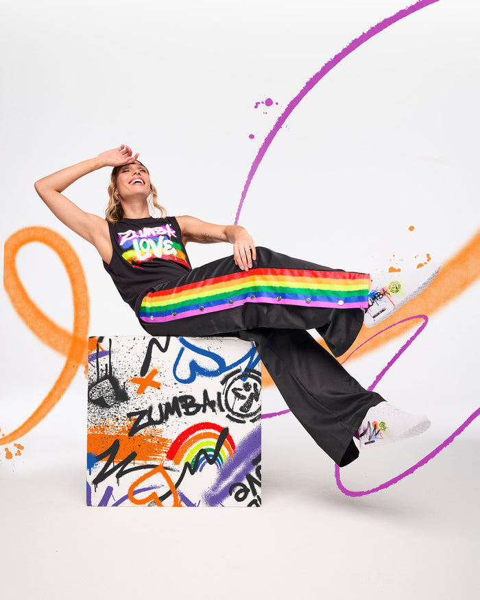 Female model wearing a black “Zumba Love” shirt with a rainbow design and black pants with a rainbow stripe down her right leg, posed against a white background with colorful paint‑style graphics.