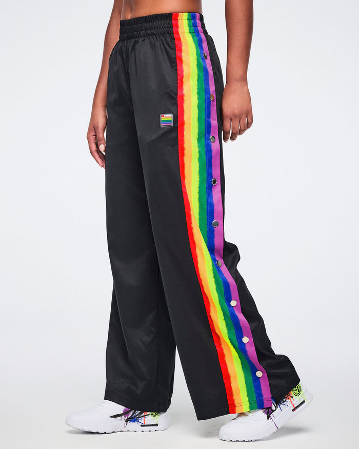 Female model wearing black track pants with rainbow side stripes and white sneakers, standing against a white background.