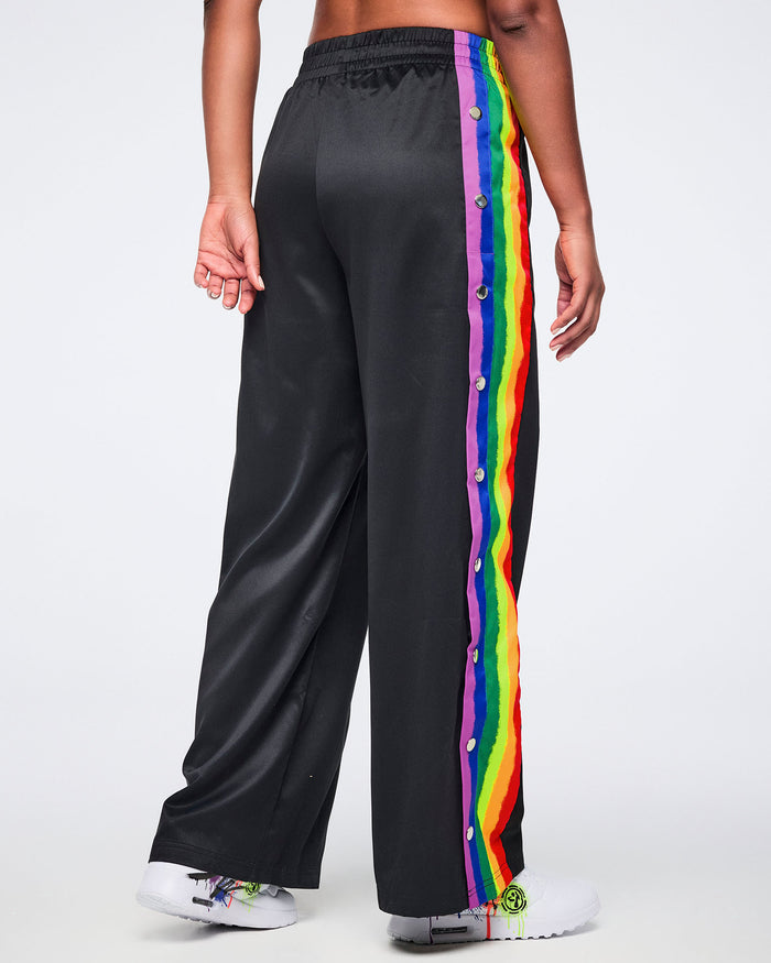 Female model wearing black track pants with rainbow side stripes and white sneakers, shown from the back against a white background.