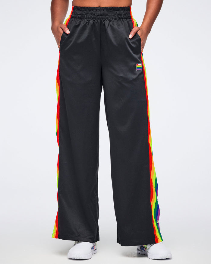 Female model wearing black track pants with rainbow side stripes and white sneakers, standing facing forward against a white background.