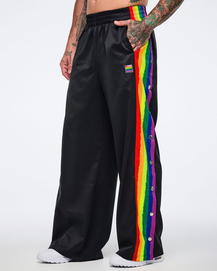 Male model in black track pants with rainbow side stripes and white sneakers, left hand in pocket, standing against a white background.