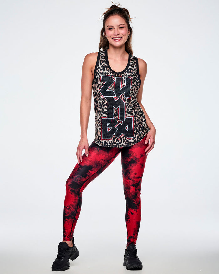 Full view of a female model wearing a basketball tank top with a black, beige, and white leopard print pattern, featuring large vertical 'ZUMBA' rock-like text in black with red and white outlines, paired with red and black leggings and black shoes.