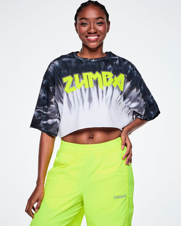 A female model wearing a cropped tie-dye Zumba top with neon yellow 'ZUMBA' rock-like text across the chest, paired with yellow pants.