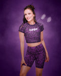 A female model wearing a matching purple and black snakeskin-patterned activewear set consisting of a short-sleeve crop top with 'ZUMBA' printed on the chest, in silver and matching high-waisted bike shorts, set against a smoky purple studio background.