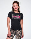 A female model wearing a black short-sleeve top with a large 'ZUMBA' text print across the chest. The letters are filled with a leopard print pattern and overlaid with a smaller pink ZUMBA font, and paired with tan leopard print biker shorts.