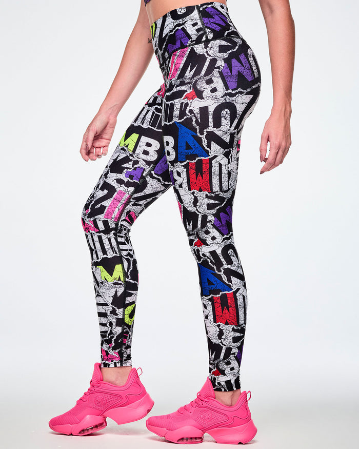 A female model wearing high-waisted leggings with a bold multicolor graffiti-style print featuring black, white, purple, blue, red, and pink lettering, paired with bright pink shoes.