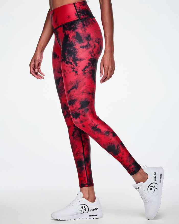 Side view of a female model wearing high-waisted red and black tie-dye like leggings, paired with white shoes with a big black Zumbito faded logo and black Zumba text.