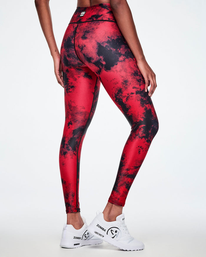 Back view of a female model wearing high-waisted red and black tie-dye like leggings, paired with white shoes with a big black Zumbito faded logo and black Zumba text.