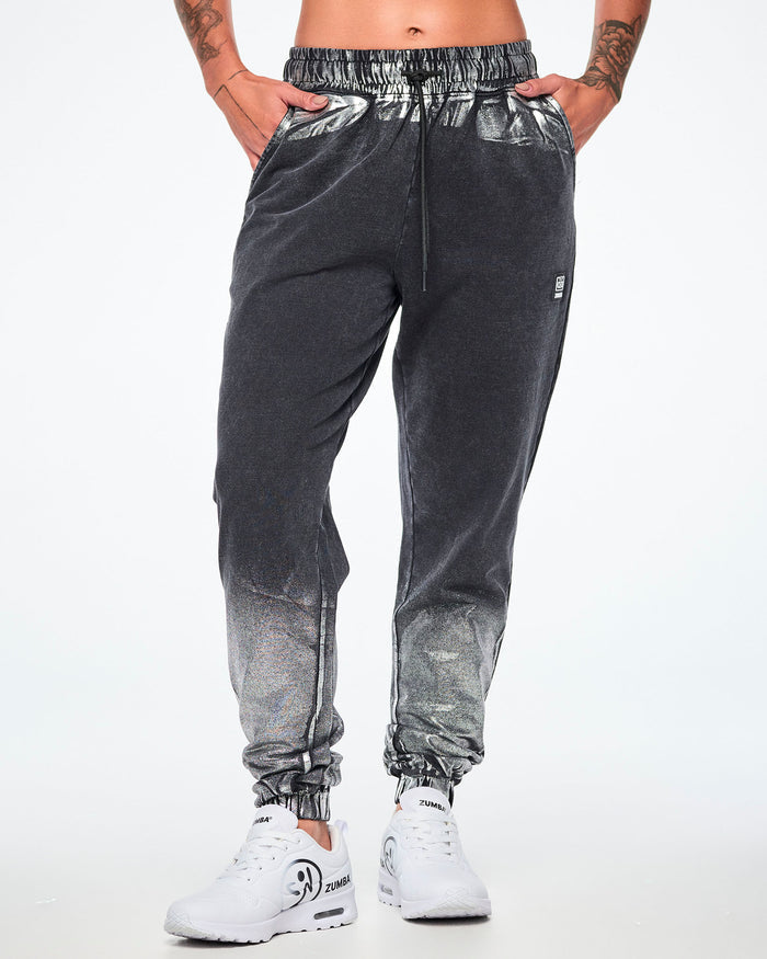 Front view of a female model wearing black and silver dark acid-wash sweatpants with a subtle tonal pattern, featuring a drawstring elastic waist, side pockets, and elastic cuffs, paired with white shoes.
