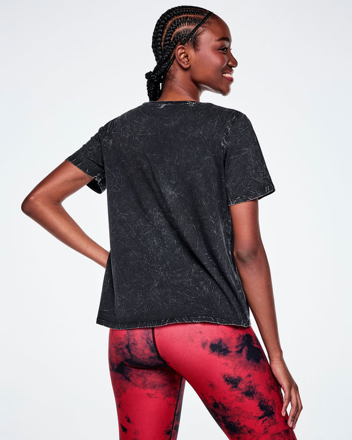 Back view of a female model wearing a black acid-washed patterned Zumba graphic tee, paired with red and black leggings.