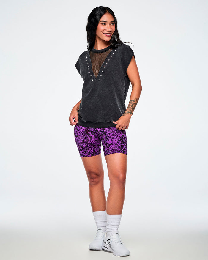 A female model wearing a acid-washed patterned black raw edge short-sleeve top with a mesh V-neckline and silver studs, paired with purple snake-skin patterned biker shorts and white shoes and socks.