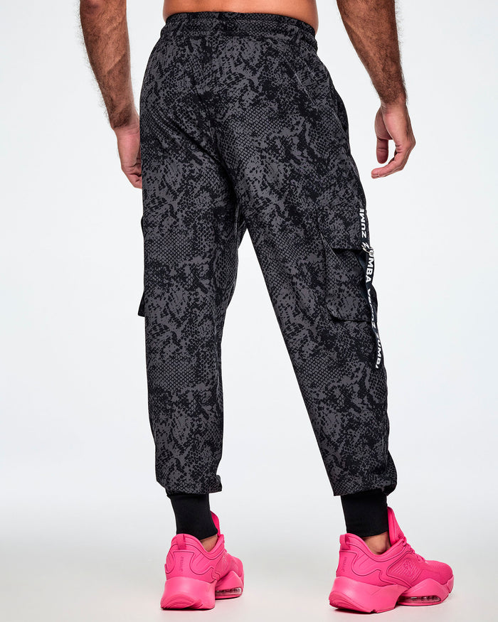 Back view of a male model wearing black and grey snake-skin patterned cargo pants with white vertical Zumba text the black straps on the cargo pockets and showing the black leg cuffs while paired with pink shoes.
