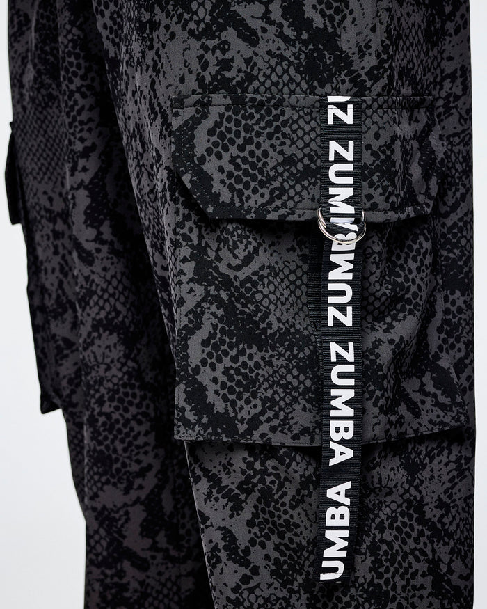 Close-up of black and grey snakeskin-patterned cargo pants with a vertical black strap on the cargo pocket with white 'ZUMBA' text.