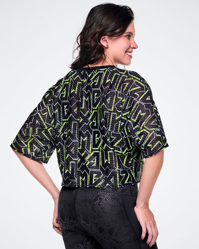 Back view of a female model wearing a short-sleeve mesh boxy crop top with rock-like letter patterning that says "ZUMBA" in black and outlined in green, and paired with black leggings.