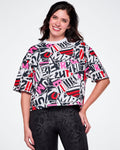 A female model wearing a short-sleeve crop top with a colorful abstract ZUMBA text pattern in pink, red, black and white, paired with black leggings.