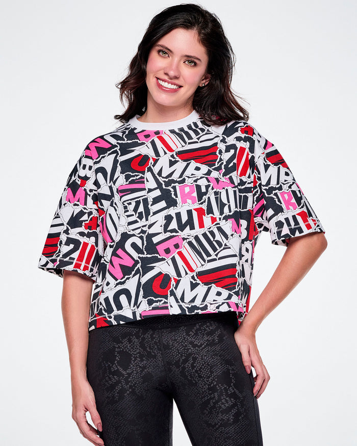 A female model wearing a short-sleeve crop top with a colorful abstract ZUMBA text pattern in pink, red, black and white, paired with black leggings.