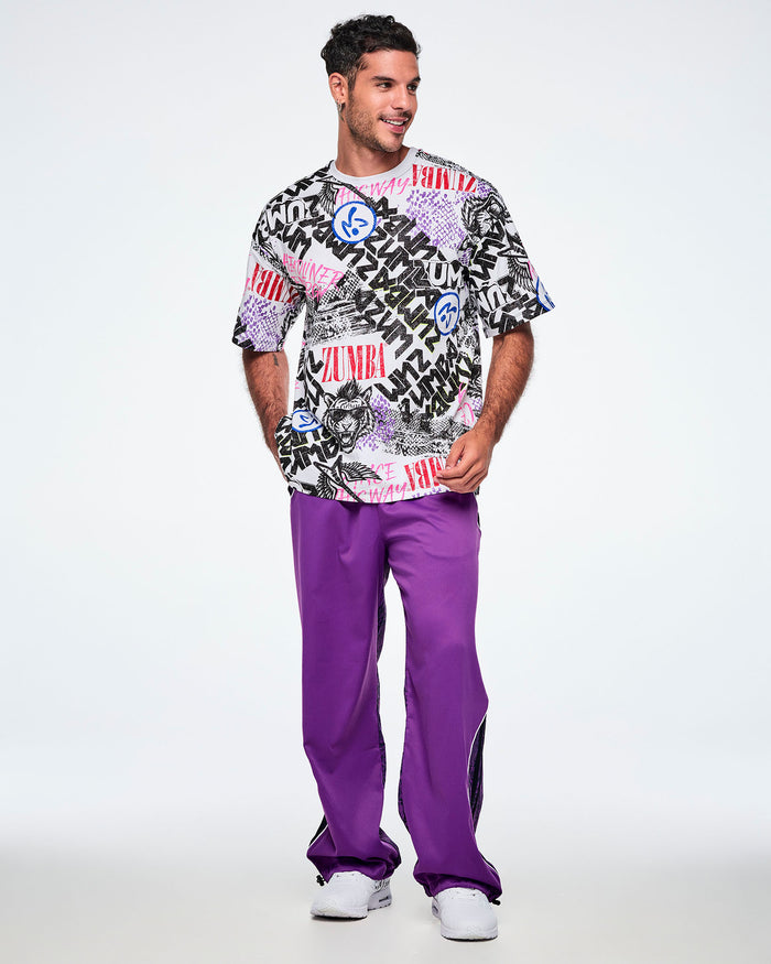 Full view of a male model wearing a graphic tee with black, white, pink, and blue graffiti-style print and logos, such as blue zumbitos, a tiger wearing sunglasses, "Headliner energy, "Dance this Way," and ZUMBA in various rock and metal text, paired with purple pants and white shoes.