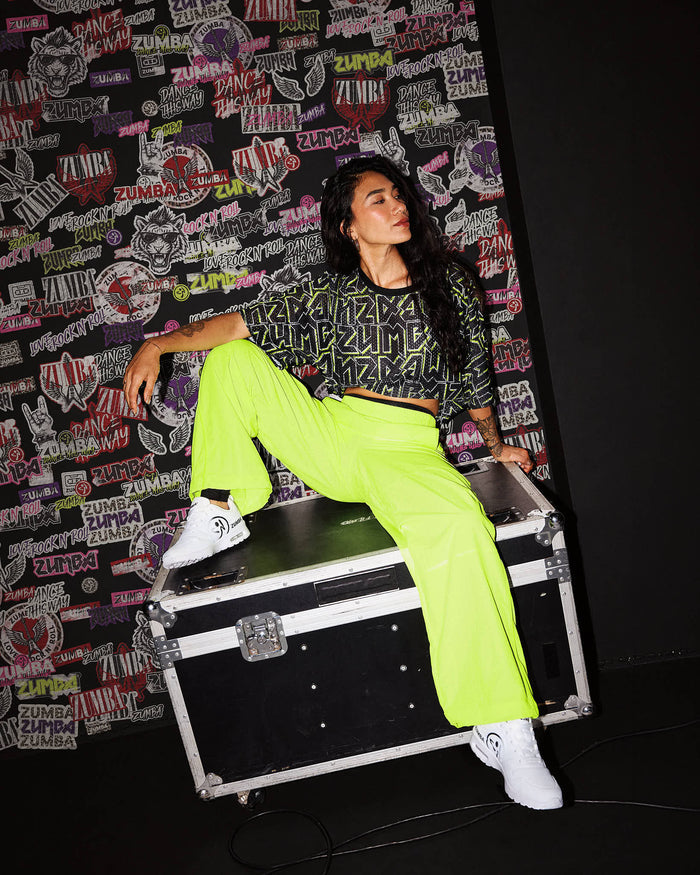 A female model wearing yellow-green pants and a matching black and yellow-green ZUMBA crop top who sits on an equipment case in front of a wall covered in Zumba rock emblems.