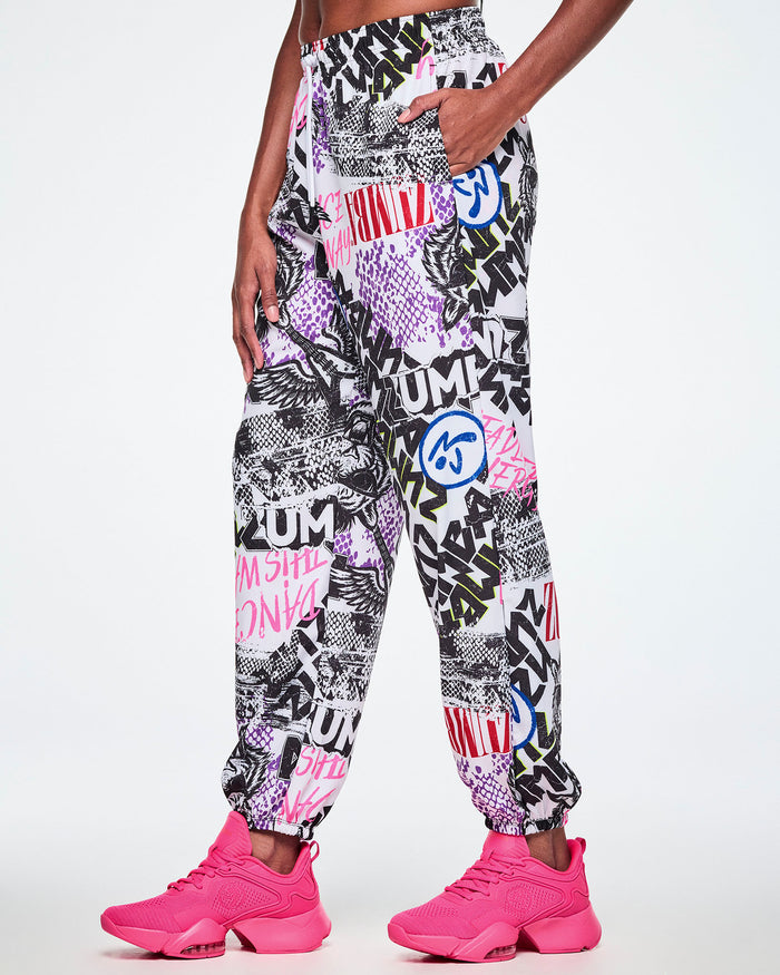 A female model wearing white woven pants with an all-over ripped newspaper like pattern with black, white and red ZUMBA text and pink Dance this way text along with other Zumba rock graphics, Zumbitos and purple and black snakeskin patterns, paired with pink shoes.