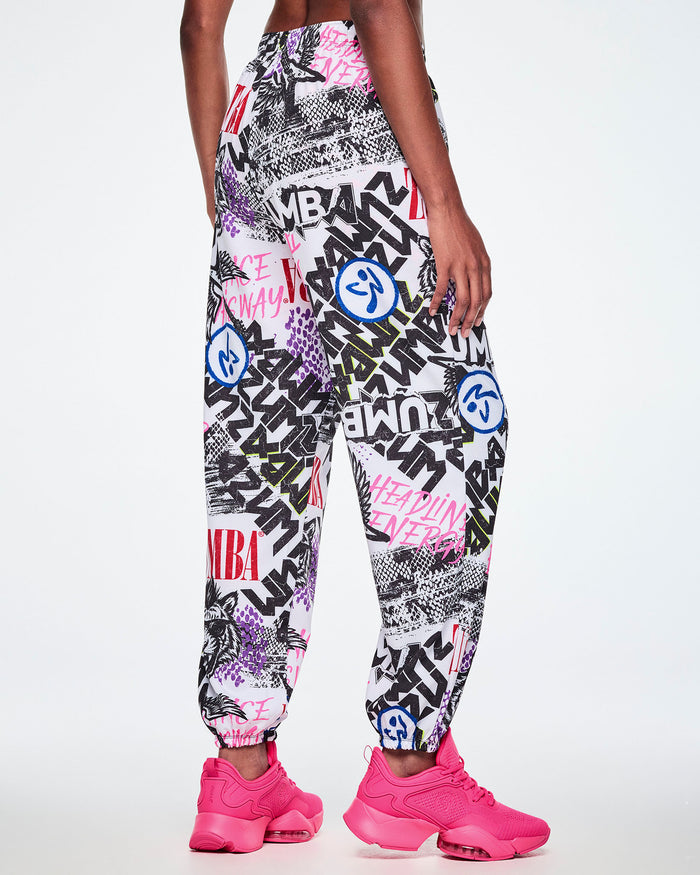 Back view of a female model wearing white woven pants with an all-over ripped newspaper like pattern with black, white and red ZUMBA text and pink Dance this way text along with other Zumba rock graphics, Zumbitos and purple and black snakeskin patterns, paired with pink shoes.