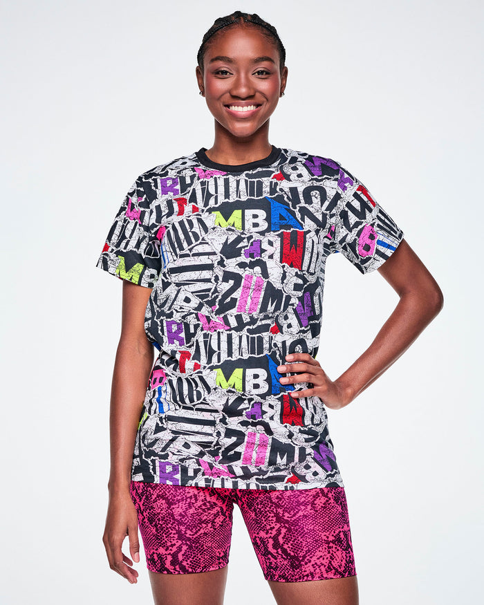 A female model wearing a multi-colored ZUMBA graphic tee and pink snake-skin patterned shorts.