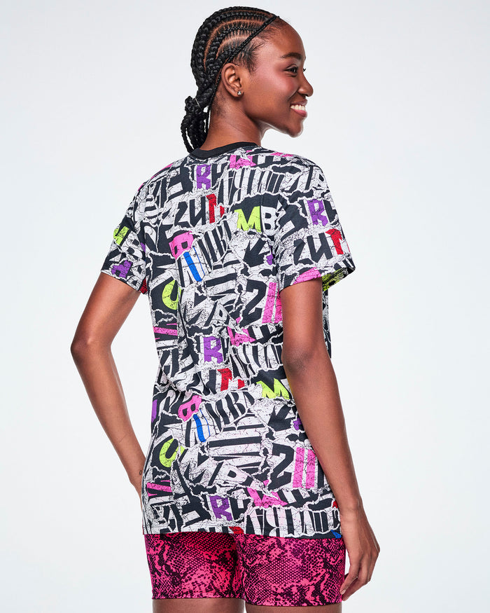Back view of a female model wearing a multi-colored ZUMBA graphic tee and pink snake-skin patterned shorts.