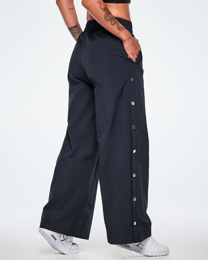 Back side view of a female model wearing wide-leg black pants showing the decorative silver-tone snap buttons down the outer side of the legs and paired with white shoes.