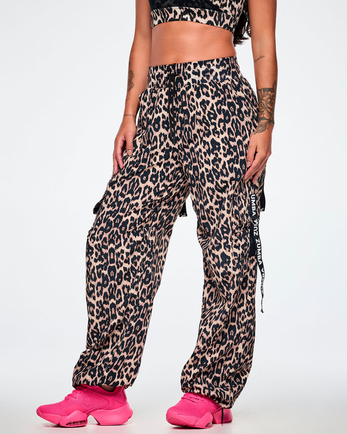 A female model wearing tan leopard print cargo pants with black Zumba straps on the pockets, paired with bright pink sneakers against a plain white studio background.