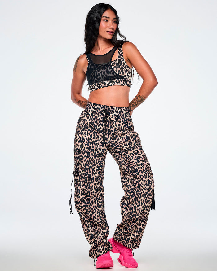 A female model wearing a two-piece outfit consisting of a black asymmetrical mesh and leopard print crop top with matching leopard print cargo pants, paired with bright pink sneakers.
