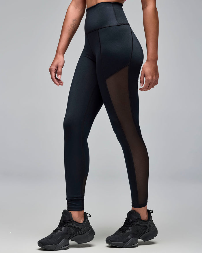 Black high-waisted leggings with sheer mesh side panels and a sleek fitted shape. Female model wears the full-length leggings with black athletic sneakers in a side view against a light gray background.