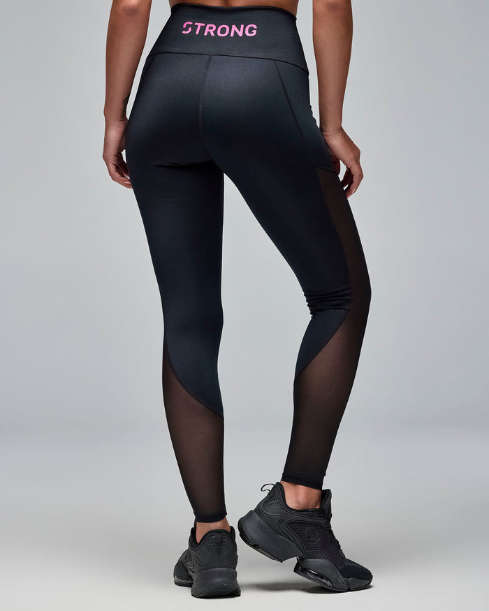 Back view of black high-waisted leggings with sheer mesh side panels and pink “STRONG” text on the waistband. Female model wears the full-length leggings with black athletic sneakers against a light gray background.