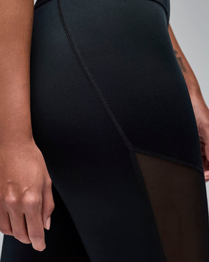 Black high-waisted leggings with sheer mesh side panels in a curved diagonal design. Female model wears the fitted full-length leggings in a close-up view against a light gray background.