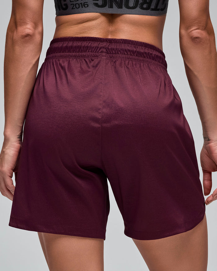 Close-up back view of burgundy athletic shorts with an elastic waistband and smooth relaxed silhouette. Female model wears the shorts against a light gray background.