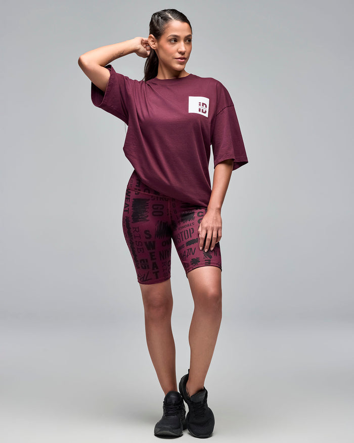 Burgundy oversized short-sleeve T-shirt with a small white chest logo patch. Female model wears the relaxed tee with burgundy printed biker shorts and black sneakers, standing with one hand behind her head against a light gray background.