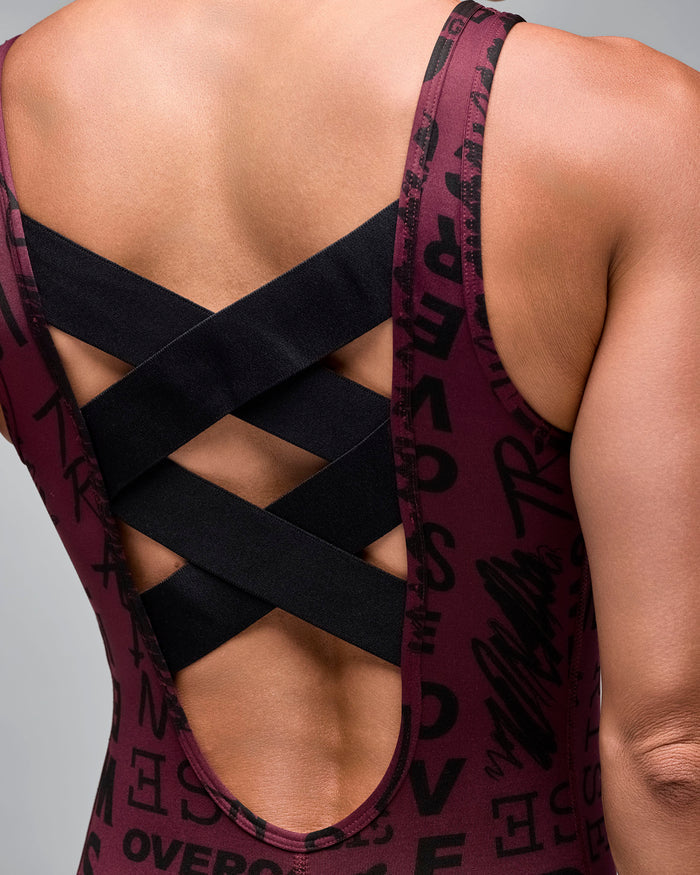 Close-up back view of a burgundy printed bodysuit featuring black crisscross straps over a low scoop back. Female model wears the sleeveless one-piece against a light gray background.