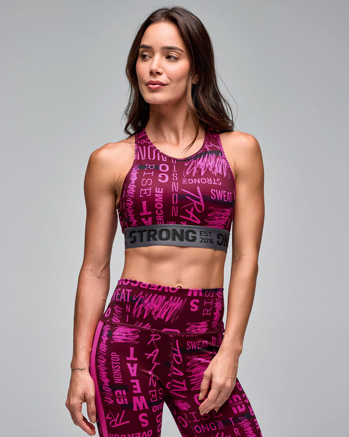 Burgundy and pink graphic-print sports bra with a gray elastic under-band, shown closely on a female model wearing matching high-waisted leggings in a front view against a light gray studio background.