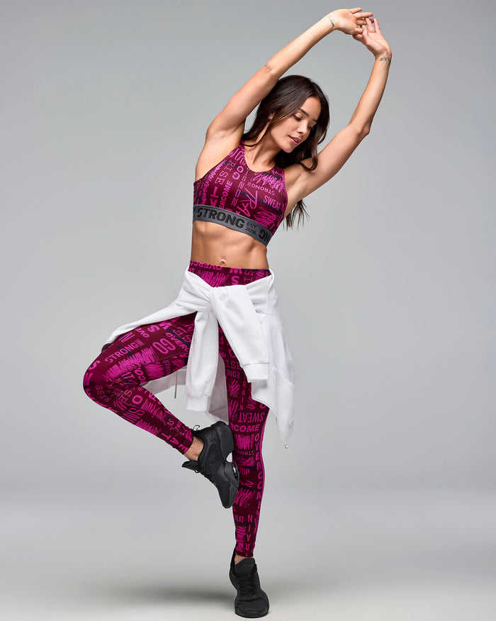 Burgundy and pink graphic-print sports bra with a gray elastic under-band, shown on a female model wearing matching high-waisted leggings, a white hoodie tied around the waist, and black sneakers in a dynamic pose.