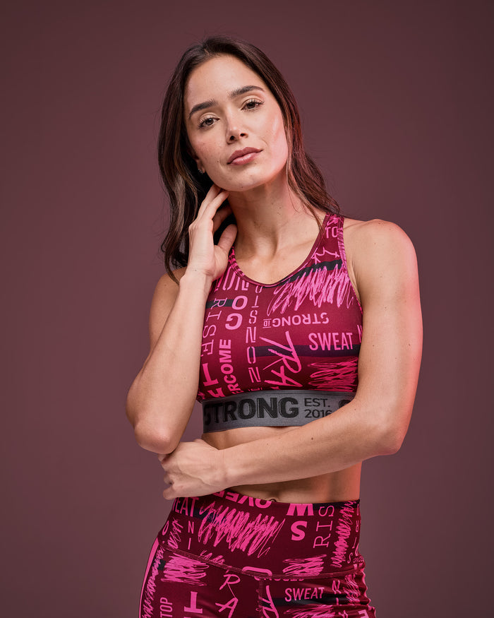 Burgundy and pink graphic-print sports bra with a gray elastic under-band, shown on a female model in a close-up front view with matching high-waisted leggings against a dark mauve studio background.