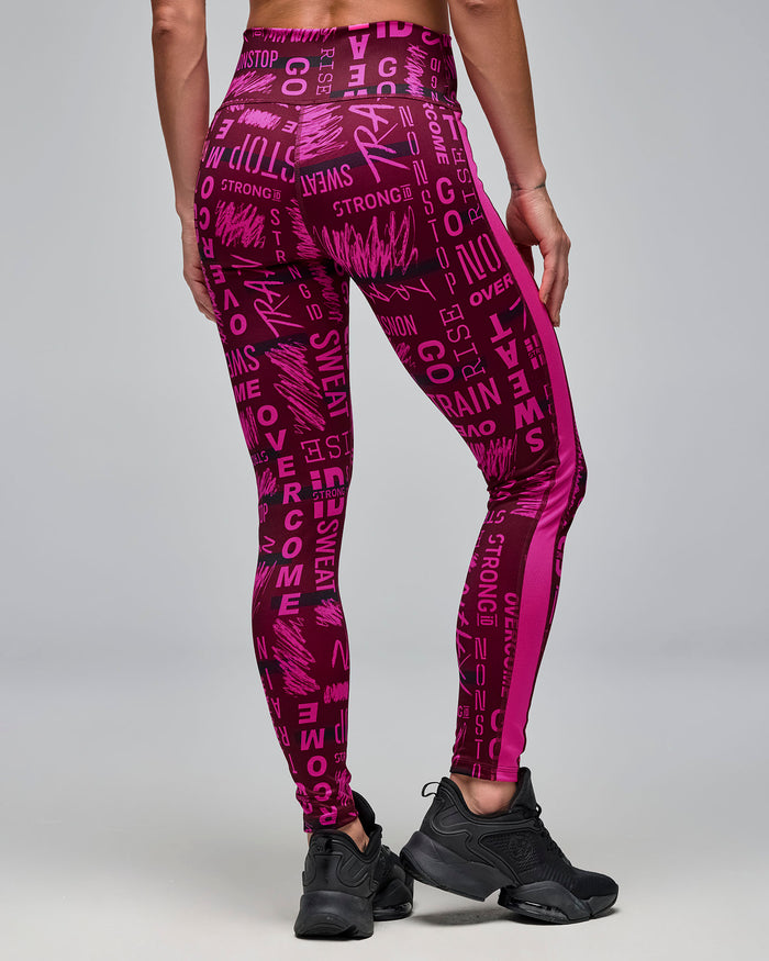 Back view of burgundy and pink high-waisted ankle leggings with an allover motivational word print and bright pink side panels. Female model wears the fitted leggings with black athletic sneakers against a light gray background.