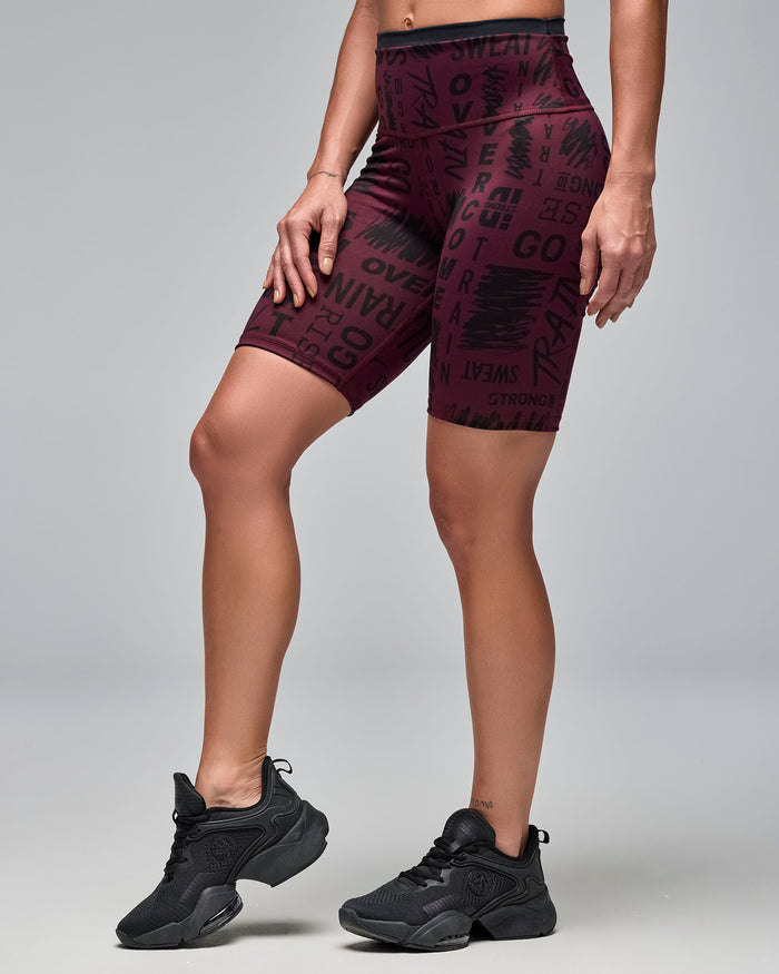 Burgundy high-waisted biker shorts with an allover black motivational word print. Female model wears the fitted mid-thigh shorts with black athletic sneakers in a side view against a light gray background.