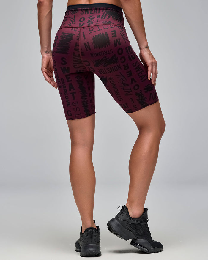 Back view of burgundy high-waisted biker shorts with an allover black motivational word print. Female model wears the fitted mid-thigh shorts with black athletic sneakers against a light gray background.