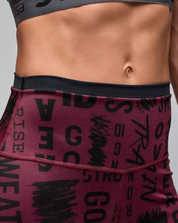 Female model wears the fitted shorts in a close-up waist view against a light gray background. She wears a burgundy high-waisted biker shorts with an allover black motivational word print and a black waistband trim.
