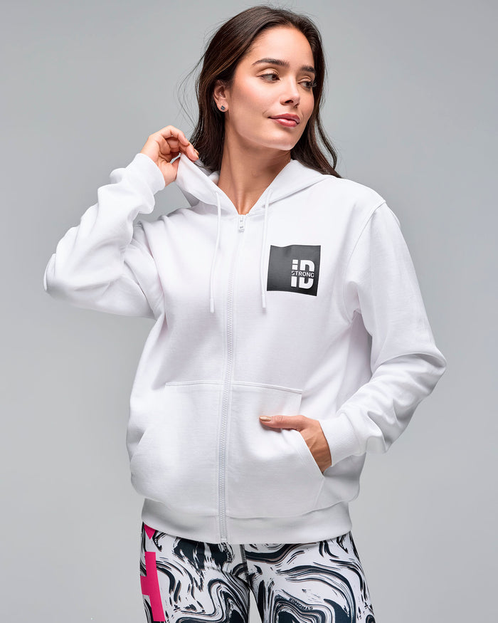White zip-up hoodie with drawstrings, front pockets, and a black chest logo patch. Female model wears the long-sleeve hoodie zipped up over white and black swirl-print leggings against a light gray background.