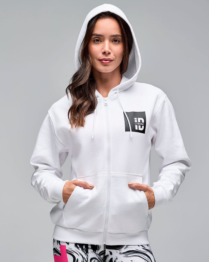 White zip-up hoodie with drawstrings, front pockets, and a black chest logo patch. Female model wears the long-sleeve hoodie with the hood up over white and black swirl-print leggings against a light gray background.