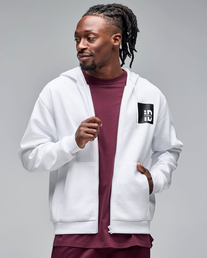 White zip-up hoodie with drawstrings and a black chest logo patch. Male model wears the long-sleeve hoodie open over a burgundy crewneck top against a light gray background.