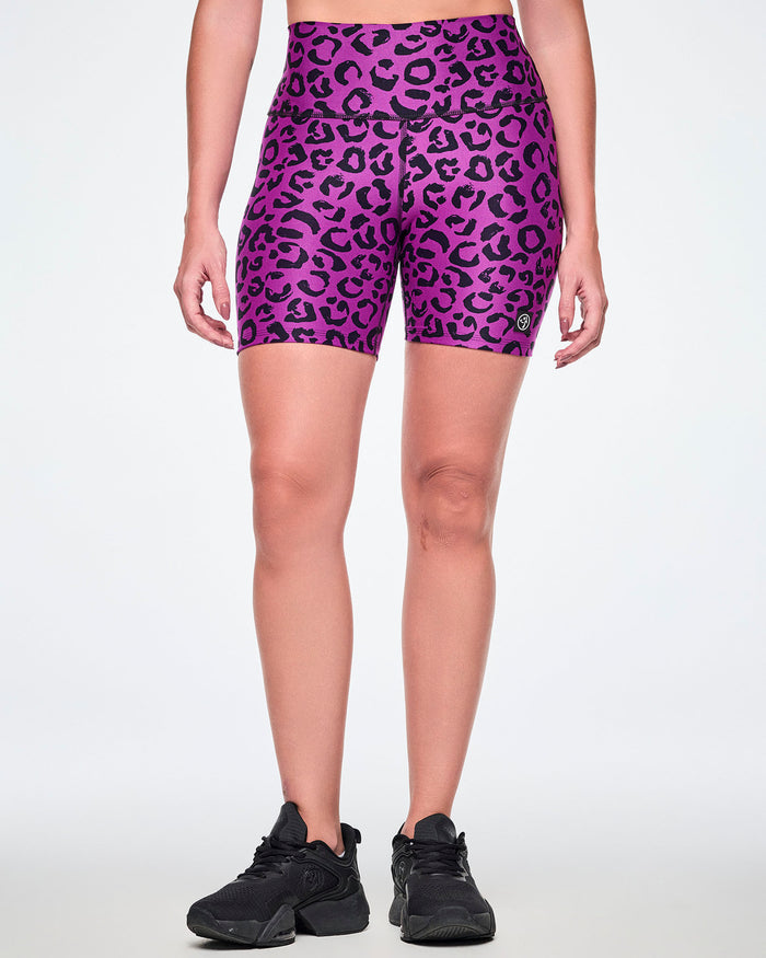 Front view of a female model wearing high-waisted purple biker shorts with a black leopard print pattern and a small Zumbito logo near the hem and paired with black shoes.