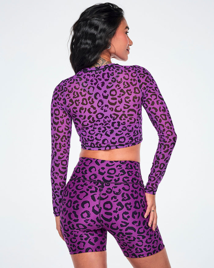 Back view of a female model wearing a matching purple and black leopard-print activewear set, featuring a long-sleeve mesh crop top and high-waisted biker shorts, against a white background.